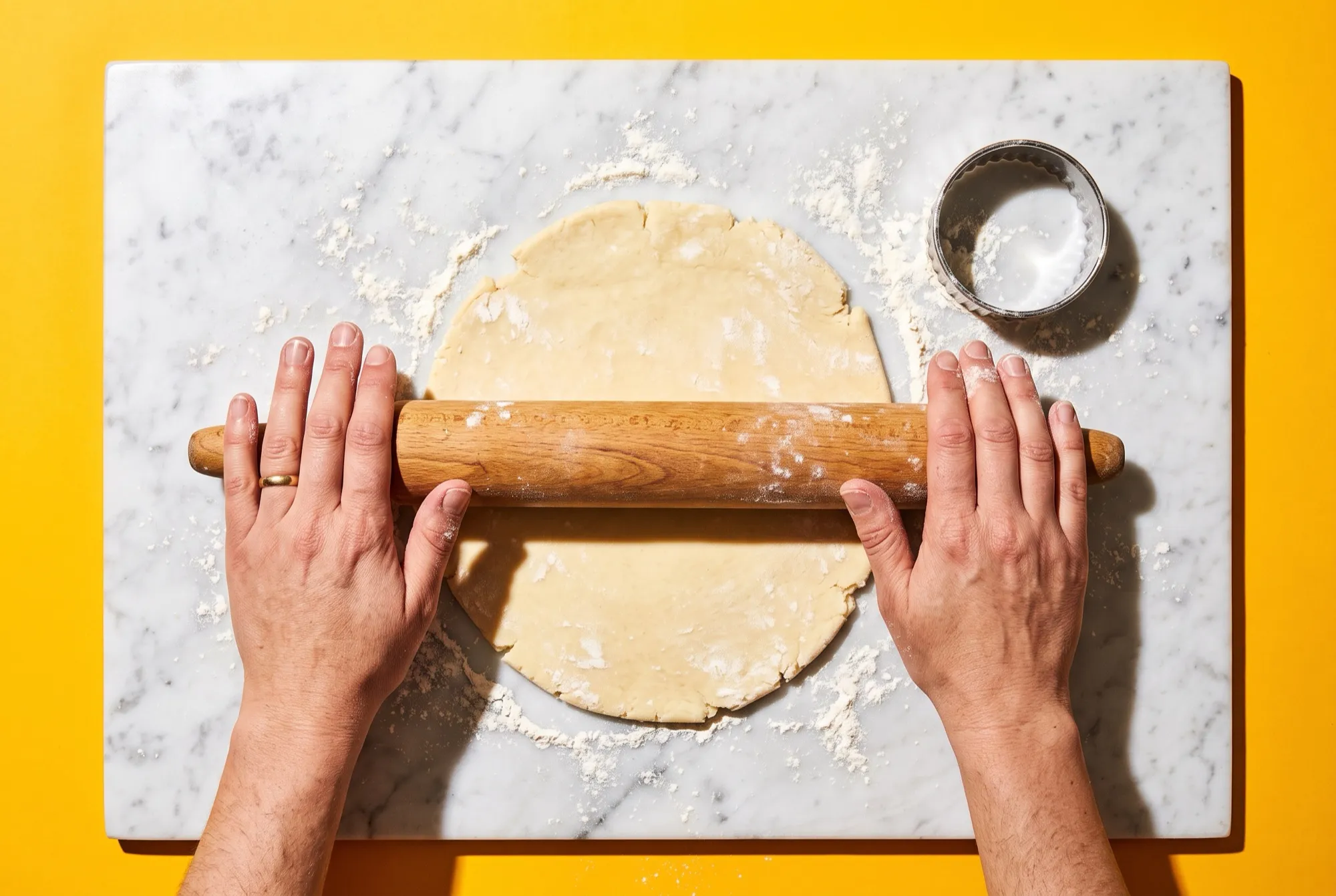 Hands rolling out butter pastry dough with a wooden rolling pin on a floured surface